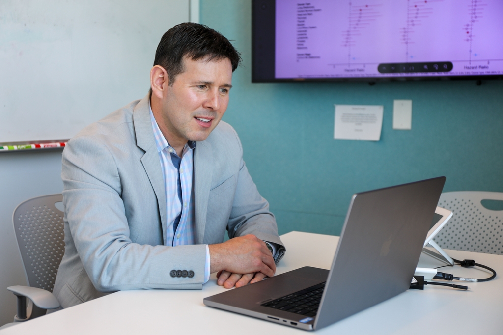 Donald sits at a desk using a laptop for a virtual conversation or presentation in an office setting, with a wall-mounted screen behind him displaying charts and data.
